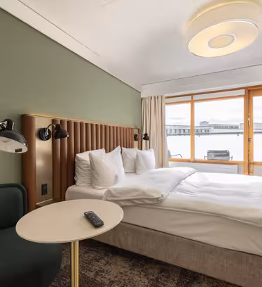 Ett hotellrum med en snyggt bäddad dubbelsäng. Rummet har en grön accentvägg och ett runt bord med en TV-fjärrkontroll. En plyschstol sitter i närheten. Stora fönster ger naturligt ljus och utsikt över stadsbilden.