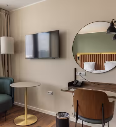 Hotellrum med ett litet skrivbord och stol, en rund spegel ovanför och en lampa fäst vid den. En platt-TV hänger på väggen. Ett runt bord med en grön stol står vid fönstret, med gardiner hängande bredvid. En papperskorg ligger på golvet.