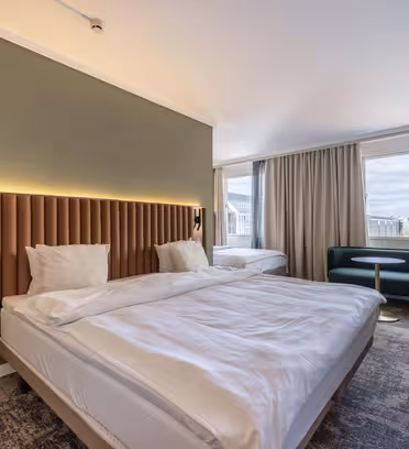 Ett modernt hotellrum med en stor, prydligt bäddad säng med en tuftad sänggavel mot en grön vägg. Ett fönster med beiga gardiner avslöjar en stadsutsikt. Rummet har en liten sittgrupp med en grön soffa och ett skrivbord med en lampa.