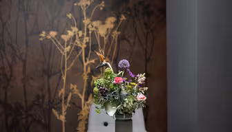 En vas med en blandad bukett av blommor, inklusive rosa, lila och gröna sorter, placeras på ett bord. Bakgrunden har en mörk vägg med ett gyllene blommönster.