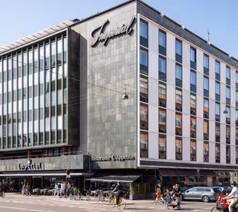 Stor byggnad med modern fasad och en skylt med texten Imperial. Bottenvåningen rymmer en restaurang med uteservering. Folk cyklar och går längs gatan. Flera parkerade bilar syns framför. Klarblå himmel i bakgrunden.