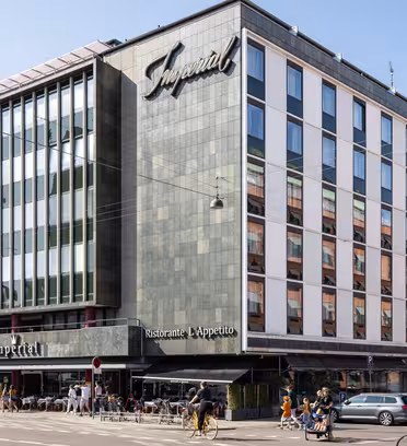 Stor byggnad med modern fasad och en skylt med texten Imperial. Bottenvåningen rymmer en restaurang med uteservering. Folk cyklar och går längs gatan. Flera parkerade bilar syns framför. Klarblå himmel i bakgrunden.