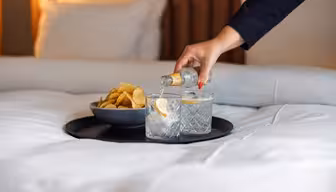 En hand häller en klar dryck från en liten flaska i ett glas med is och citron, bredvid ett annat glas och en skål med potatischips på en bricka ovanpå en prydligt bäddad säng.