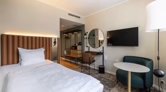 Ett modernt hotellrum med enkelsäng, stoppad sänggavel, vägghängd TV, rund spegel, skrivbord med stol, grön fåtölj, litet runt bord, golvlampa och öppen garderob. Inredningen är minimalistisk och i neutrala toner.