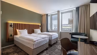 Modernt hotellrum med två enkelsängar, stoppad sänggavel, grön accentvägg, stora fönster med utsikt över staden, ett runt bord med stolar och en TV på väggen. Neutrala färger och modern inredning.