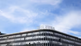 En modern kontorsbyggnad i glas med horisontella linjer under en klarblå himmel med spridda moln. Byggnaden har en skylt ovanpå med stora bokstäver och trafikljus syns i hörnet.
