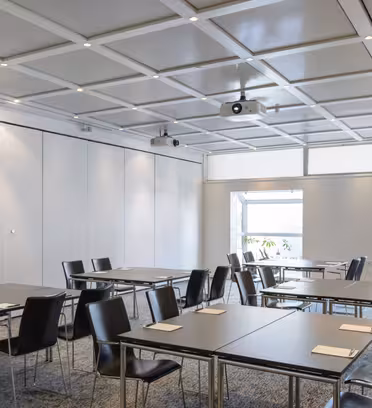 Ett modernt konferensrum med arrangerade rektangulära bord och svarta stolar. Mjuk belysning från takarmaturer och naturligt ljus från stora fönster med växter synliga, skapar en professionell och öppen atmosfär.