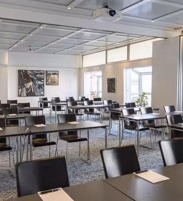 Ett konferensrum med rader av svarta stolar och bord ordnade prydligt. Anteckningsblock och pennor läggs på borden. Rummet har stora fönster, flera växter och abstrakt konst på väggarna. En takmonterad projektor syns.