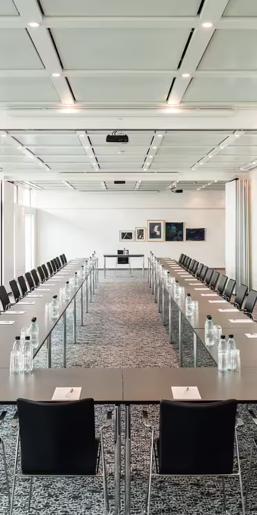 Ett rymligt konferensrum med två rader långbord som bildar en U-form. Varje bord har svarta stolar och är inrett med anteckningsblock, pennor och vattenflaskor. Rummet är väl upplyst med stora fönster och inramad konst på en avlägsen vägg.
