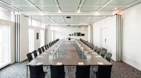 Ett rymligt konferensrum med två rader långbord som bildar en U-form. Varje bord har svarta stolar och är inrett med anteckningsblock, pennor och vattenflaskor. Rummet är väl upplyst med stora fönster och inramad konst på en avlägsen vägg.