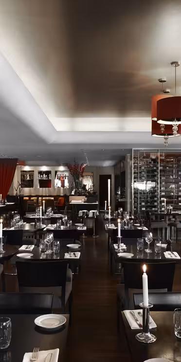 Exklusiv restauranginteriör med svag belysning, snyggt dukade bord med ljus och glas. Modern inredning inkluderar röda ljuskronor, konstverk på väggarna och ett stort vinställ. Atmosfären är elegant och inbjudande.