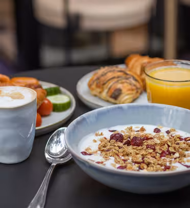 Ett frukostpålägg med en skål yoghurt med granola och torkad frukt, en tallrik med croissanter, gurka och skivade apelsiner, ett glas apelsinjuice och en kopp kaffe med skummande mjölk, allt på ett mörkt bord.