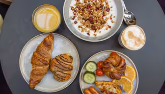 En frukost som serveras på ett bord inkluderar en skål med yoghurt med granola, ett glas apelsinjuice, en kopp cappuccino, croissanter, ett bakverk, skivade apelsiner, gurka, körsbärstomater, minikorvar och bacon.