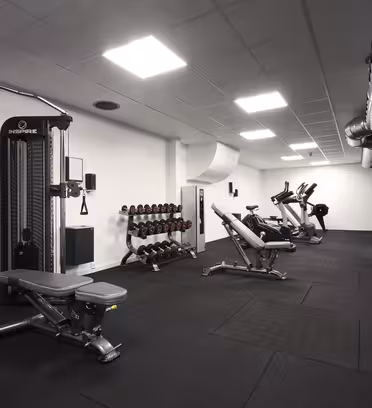 Ett modernt gym med olika träningsutrustning inklusive viktmaskiner, hantlar, bänkar, löpband och stationära cyklar. Rummet har en elegant, industriell design med svart gummigolv och synliga kanalsystem.