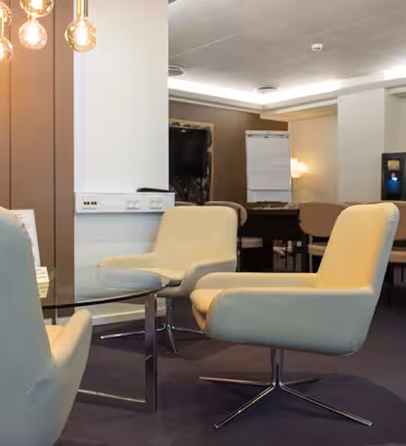 En modern mötesplats med snurrstolar runt ett glasbord. Varm takbelysning och ett neutralt färgschema skapar en mysig atmosfär. I bakgrunden finns ett blädderblock, en TV och fler sittplatser.